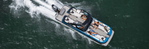 Overhead view of a motorboat moving quickly across a body of water. The boat features several seating areas, with a few people lounging and enjoying the ride under sunny conditions. Waves and water spray are visible trailing behind the boat—an exhilarating scene that might inspire you to buy pontoons.