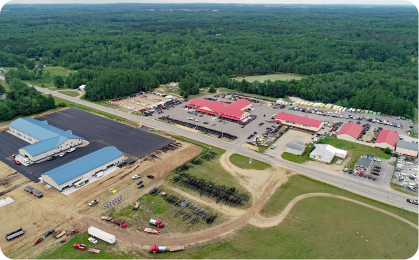 Aerial view of a large industrial area with several buildings, including blue-roofed and red-roofed structures. Surrounding the buildings are parking lots, a green forest, and open fields. Machinery and equipment are scattered around the property. A road runs through the area, where a pontoon boat for sale is visible.