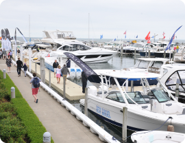 A bustling marina with numerous docked boats and yachts. People are walking along the dock, some observing the boats. Several flags and banners are visible, indicating a boat show or event. Among them is a pontoon boat for sale, attracting curious onlookers. The sky is overcast, and a landscaped path runs alongside the dock.