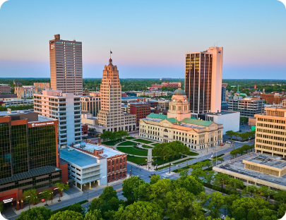 Aerial view of a cityscape at sunset featuring a mix of historic and modern buildings. A prominent courthouse with a green lawn is in the center, surrounded by several tall office buildings. Tree-lined streets and low-rise buildings are visible in the foreground, where a sign reads "Pontoon Boat for Sale.