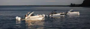 Three pontoon boats are floating on a calm body of water near a wooded shoreline under a partly cloudy sky. The boats, complete with raised sunshade canopies, appear to be stationary as people relax on each of them, likely enjoying the experience without worrying about pontoon boat prices.