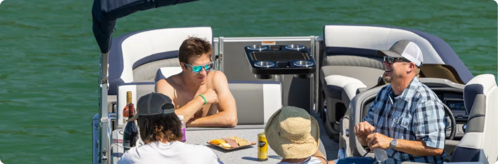 Four people are on a boat enjoying a sunny day. Three are seated, with one shirtless person wearing sunglasses at the head of the table. There are snacks and drinks on the table. The boat, fitted with used pontoons, is on calm green water, and everyone appears relaxed and cheerful.