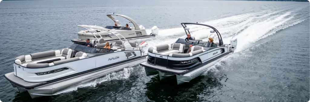 Two luxury used pontoon boats, one white and one black, speed side by side on open water. Each boat has passengers relaxing on spacious seating under sleek, curved metal canopies. The wake behind them shows their swift movement across the calm water.