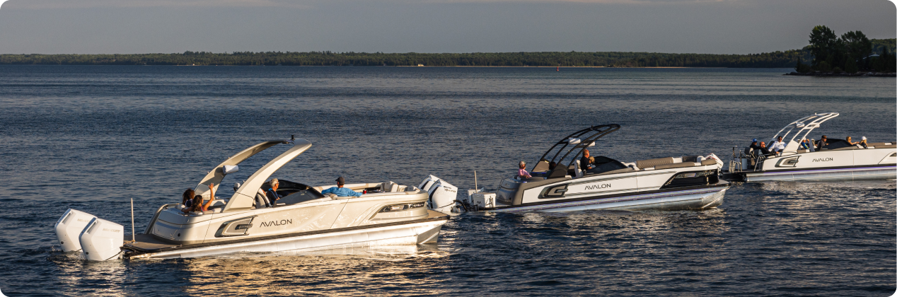 Three luxury pontoon boats with groups of people on a serene lake. The boats, which are available as a pontoon boat for sale, have sleek designs with white and silver exteriors. The lake is calm, surrounded by a forested shoreline, and the sky appears clear with soft lighting, suggesting late afternoon or early evening.