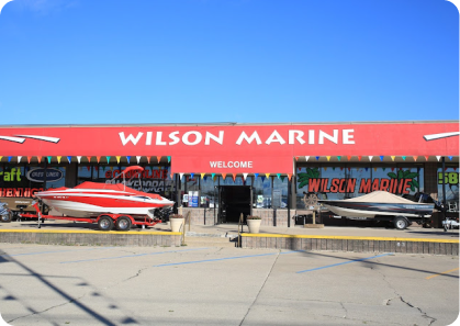 Exterior of Wilson Marine store on a clear day with a red sign reading “WILSON MARINE” and a “WELCOME” sign above the entrance. Two boats, including a pontoon boat for sale, are displayed in front of the store: one red and white, the other black and white. Multicolored flags hang above the entrance.