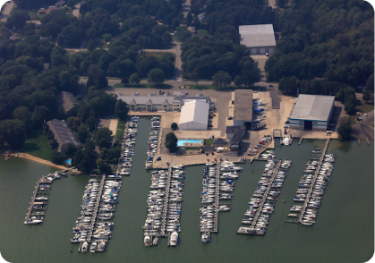 Aerial view of a marina with numerous boats docked in multiple rows extending into the water. The boats, including a pontoon boat for sale, are docked alongside several buildings and structures on land, surrounded by trees and roads. A swimming pool is visible among the buildings.