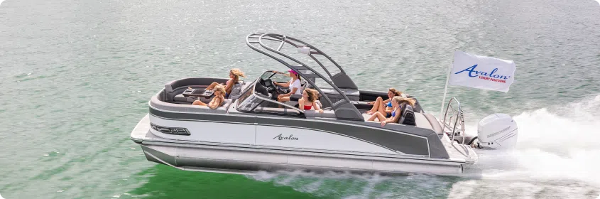 A group of people is enjoying a leisurely ride on a sleek pontoon boat labeled "Avalon" gliding through green waters. A flag with the logo "Avalon" is attached to the rear of the boat. Interested in similar experiences? Buy pontoons powered by an outboard motor, creating a wake behind them.