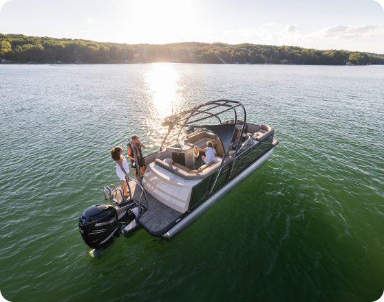 A boat with a dark hull navigates calm waters during a sunny day. Four people, two at the front and two at the rear, relax and enjoy the view. The water is green, and a forested shoreline is visible in the background, shimmering under the sunlight—an idyllic scene for those looking to buy pontoons.