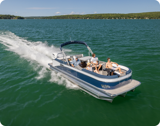 Three people are riding on a motorized pontoon boat in a large body of water surrounded by green, forested land. The boat is creating a wake as it moves, and the passengers are seated and enjoying the ride under a clear, sunny sky. It's enough to make anyone want to buy pontoons for their own adventures.