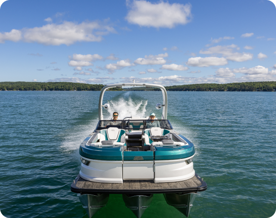 A teal and white speedboat with two people onboard speeds across a large body of water, leaving a wake behind. The sky is clear with scattered clouds, and the shoreline with trees is visible in the distance—making it a perfect day to consider why people buy pontoons for leisurely rides.