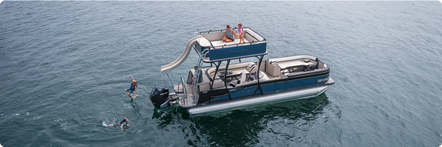 A pontoon boat with aluminum pontoons and a slide floats on a body of water. Two people in swimsuits are swimming near the boat, while two others are on the upper deck, with one preparing to use the slide. The boat is spacious with multiple seating areas.