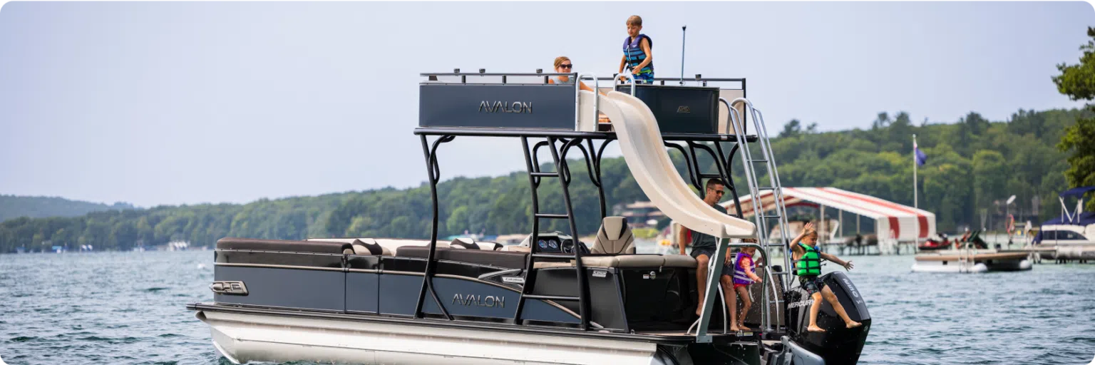 A black pontoon boat with aluminum pontoons is anchored in a lake, featuring a slide from its upper deck. Four children in life jackets are seen enjoying the boat; two on the upper deck and two using the slide. A shoreline with trees and a few buildings is visible in the background.