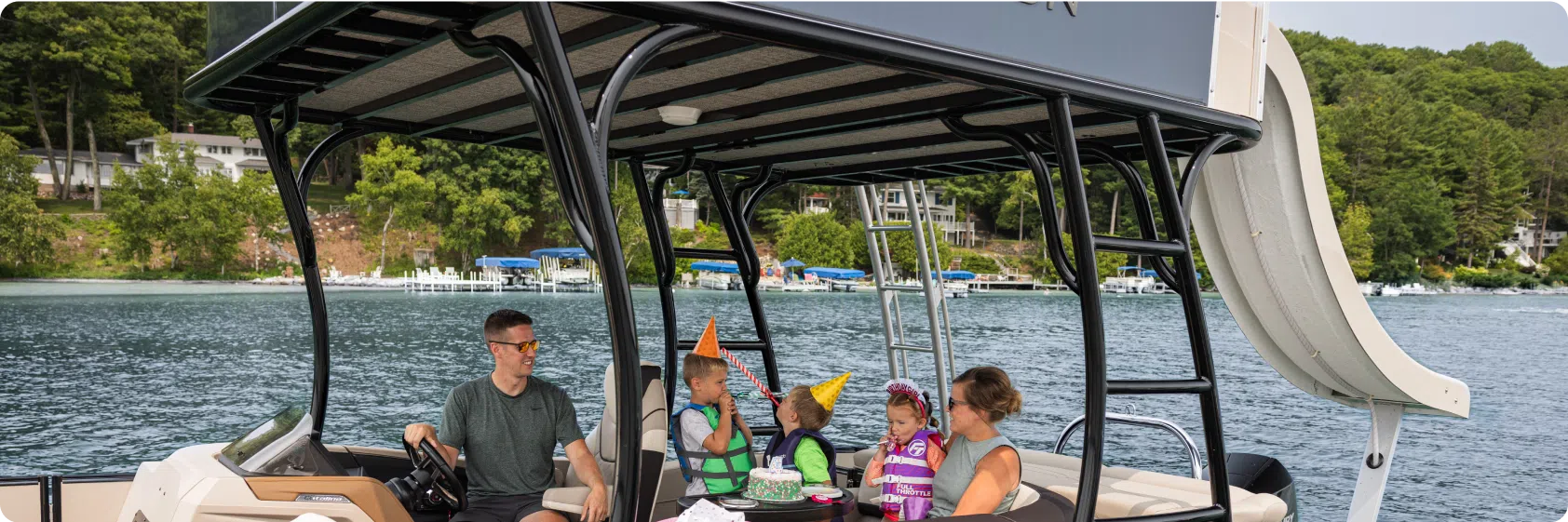 A family is celebrating a birthday on a pontoon boat with sleek aluminum pontoons. An adult steers the boat while another adult and two children in birthday hats sit around a table with a cake. The lake is surrounded by trees and other boats in the background, creating the perfect festive backdrop.