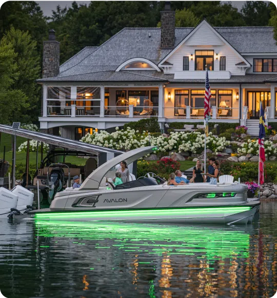 A modern boat with green underglow lights floats on a calm lake, its aluminum pontoons glistening beneath the surface. In front of a large, illuminated house with multiple flags, several people enjoy the evening, while the lush greenery and well-lit house create a serene, picturesque backdrop.