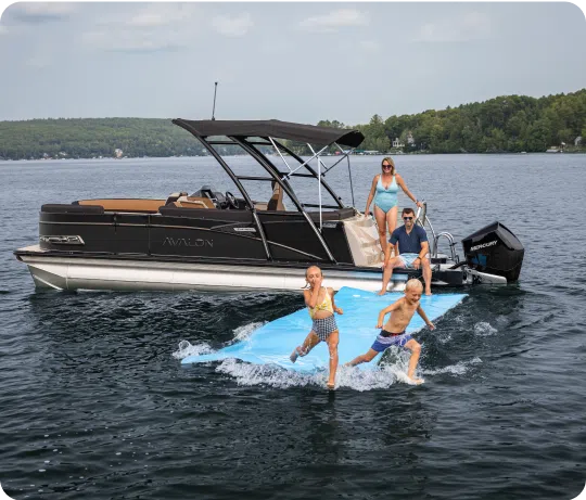 A family enjoys a day on the lake with their boat. Two children run on a floating mat in the water, while a man and woman, wearing swimsuits, watch from the boat. The "Avalon," with its aluminum pontoons gleaming in the sunlight, sits on a calm lake surrounded by trees.