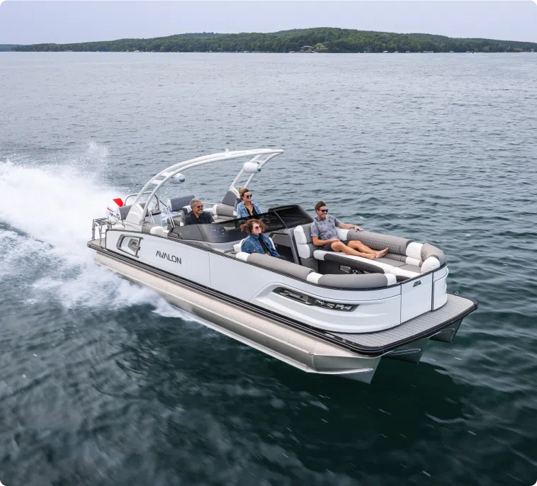 A sleek Avalon pontoon boat glides gracefully on the lake, carrying four passengers who seem relaxed and enjoying the ride. The backdrop of a forested shoreline under a clear sky completes the serene scene. With its elegance and comfort, it’s easy to see why people are curious about pontoon boat prices.