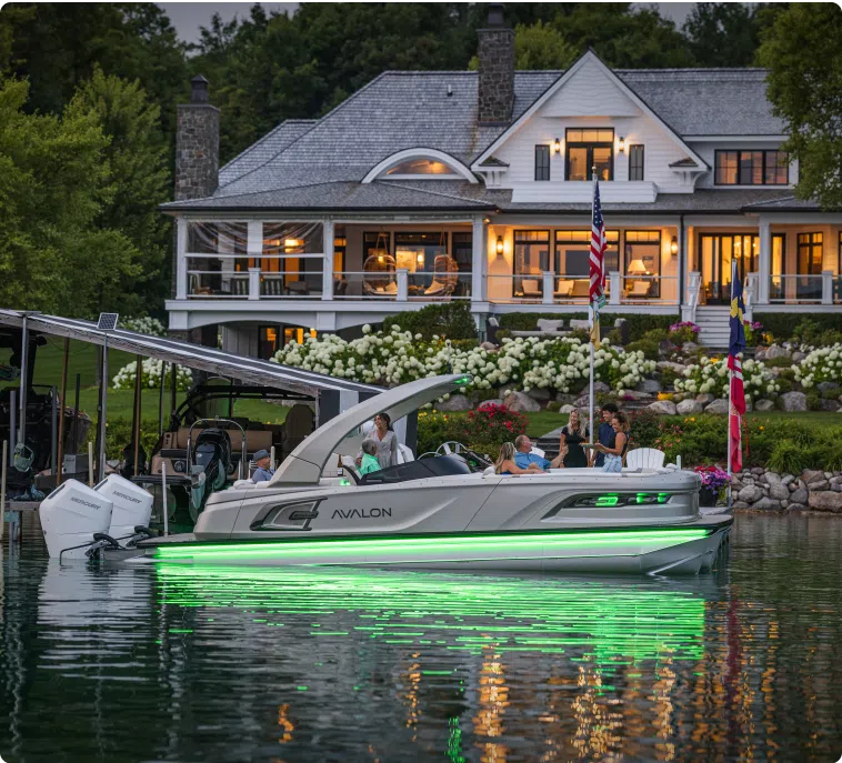 A pontoon boat with green underwater lights is docked at a lakeside house. The house has large windows and a porch with light glowing from inside. Several people are on the boat, enjoying the evening, discussing the affordable pontoon boat price, with lush greenery in the background.