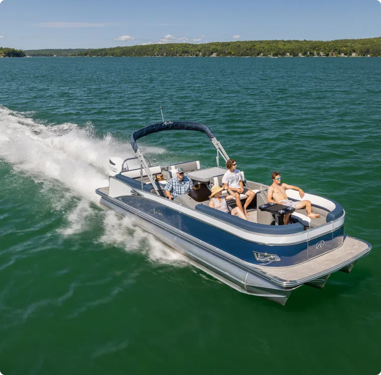 A pontoon boat with blue accents is cruising on a clear, green lake. Four people are on board; two are seated at the front, one is at the helm, and another is walking. The boat leaves a foamy white wake as it moves through the water, with a wooded shoreline visible in the distance—a scene worth every penny of the pontoon boat price.