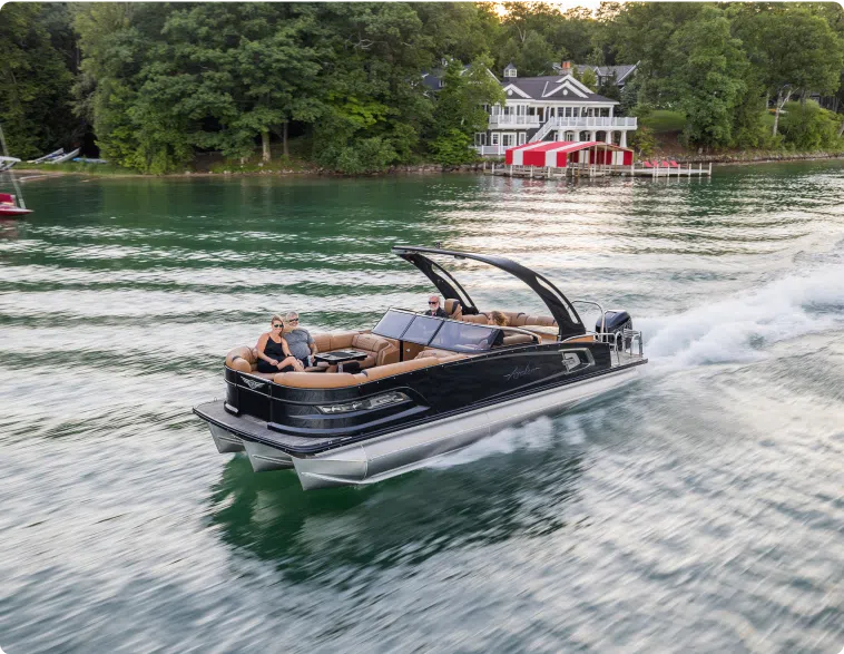 Two people are seated on a sleek, black pontoon boat cruising on a calm lake. The boat leaves a trail in the water as it moves. Lush green trees and a large house with a red and white dock are visible in the background. With such scenic views, it's easy to see why the pontoon boat price is worth every penny.