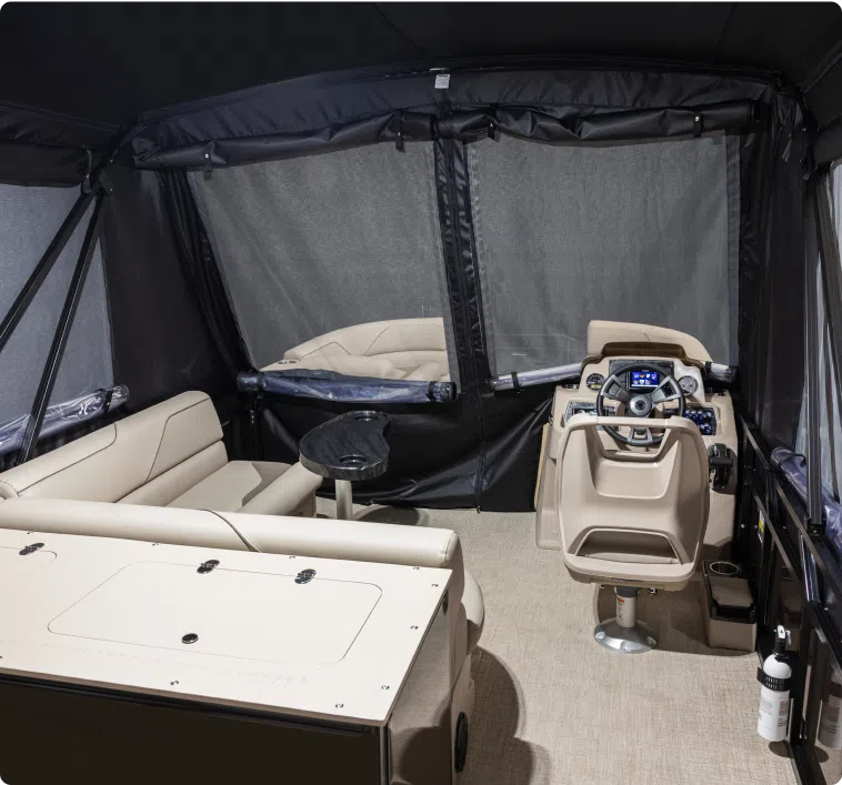 Interior view of a recreational boat, featuring beige cushioned seating, a captain's chair with steering controls, various instrumentation on the panel, and netted windows with dark curtains. The carpeted floor complements the small round table positioned in front of the seats—an elegant setup often reflective of a competitive pontoon boat price.