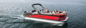 A red and black pontoon boat labeled "TAHOE" cruises on a calm body of water, showcasing the appeal of new pontoon boat sales. Five people are onboard; four are sitting and relaxing on the cushioned seats, while one steers. The sun is shining, creating a peaceful, leisurely atmosphere.