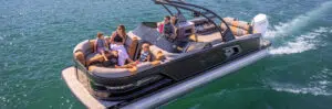 A family relaxes on a modern pontoon boat from a trusted pontoon boat dealer, cruising over a clear, greenish-blue lake. The boat features cushioned seating, a steering console, and a powerful outboard motor. The family members, including children, are enjoying the sunny weather and taking in the views.