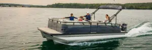 A family of four enjoys lounging on a black pontoon boat cruising on a lake. Trees and buildings are visible in the background, reflecting in the calm water. The boat, fresh from new pontoon boat sales, has a canopy providing shade for the passengers.