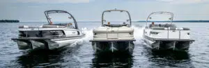 Three pontoon boats, each with two outboard engines, glide on a calm lake. The middle boat is slightly ahead. The water is relatively still, and a forested shoreline is visible in the background under a cloudy sky. Many admire the serene scene without considering the cost of pontoon boats involved.