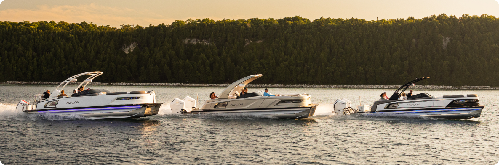 Three new pontoon boats are cruising side by side on a calm body of water at sunset, leaving behind white wakes. The boats, lit by the golden sunlight, carry passengers enjoying the ride. A tree-covered shoreline is visible in the background.