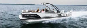 A sleek, black and white Avalon pontoon boat speeds across a calm lake. Four people are relaxing onboard, some lounging on the cushioned seats. The water is slightly choppy, and a forested shoreline is visible in the distance, illustrating why many believe the cost of a pontoon boat is worth it.