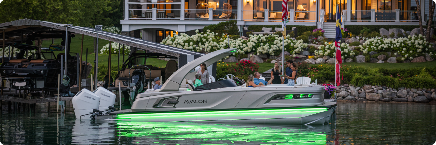 A luxury pontoon boat from a renowned pontoon boat dealer, adorned with glowing green lights, is docked near a lakeside house with a well-maintained garden. Several people are on the boat, and the house porch is lit with a warm glow. The scene is calm and inviting, reflecting beautifully off the water.