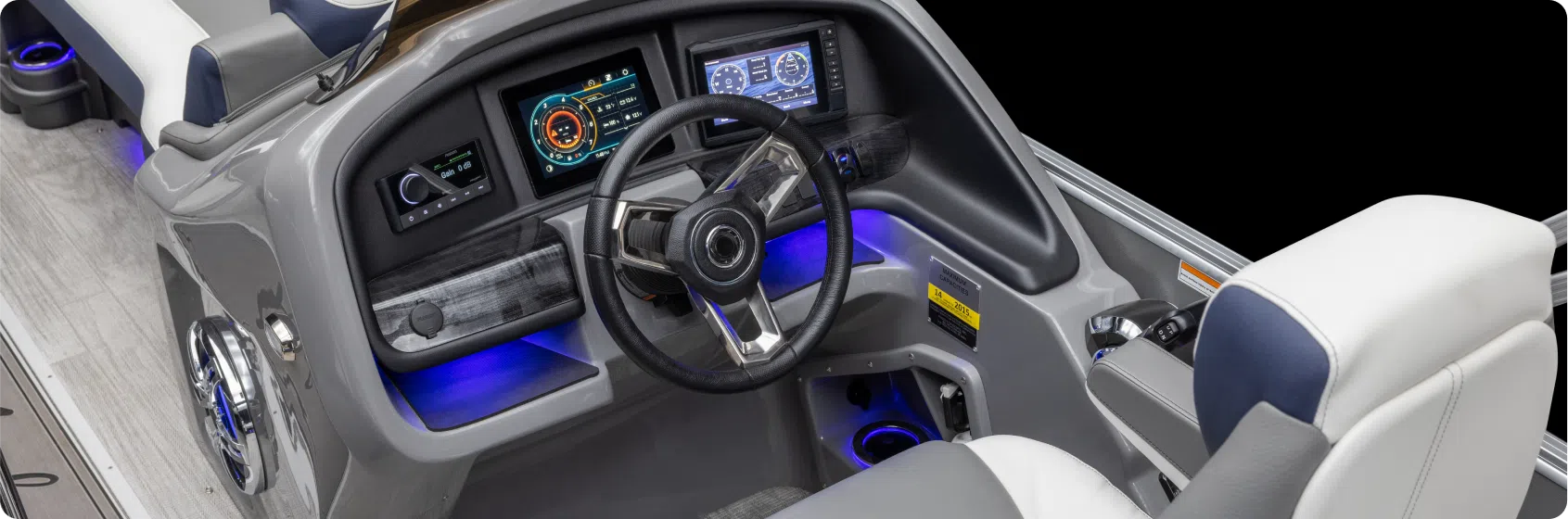 The image shows the modern cockpit of a boat, featuring a sleek steering wheel, multiple digital displays, and LED lighting accents. The cockpit area is designed with gray and white tones; the displays show various navigational and operational information you'd expect from a top pontoon boat dealer.