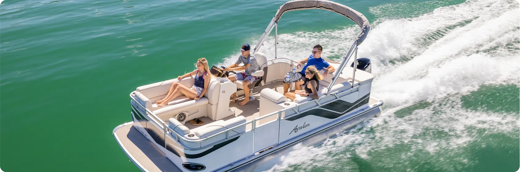 A pontoon boat from a renowned pontoon boat dealer navigates through calm, emerald-green waters. Four people are aboard; two are seated and relaxed, while the other two, including the driver, are standing. The boat creates a gentle wake as it moves forward, under sunny, clear skies.