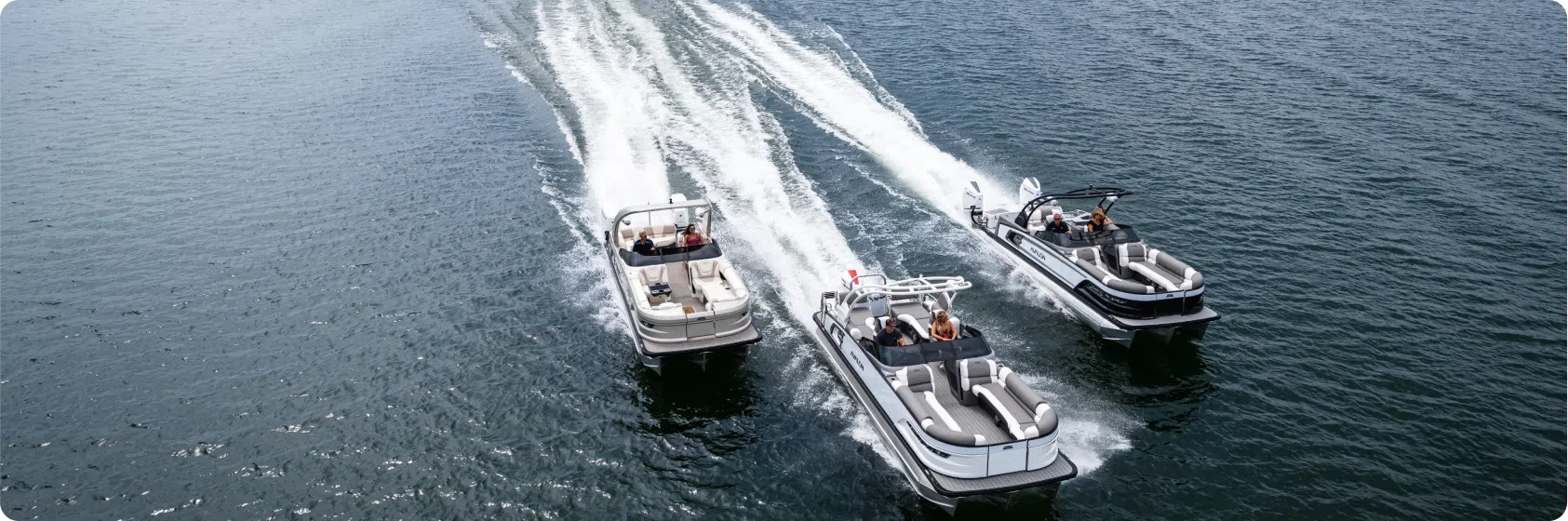 Aerial view of three motorboats cruising on a body of water, leaving white trails behind. The boats are arranged side by side with people on board each vessel, enjoying the ride under clear skies, surely a scene that would appeal to any pontoon boat dealer.