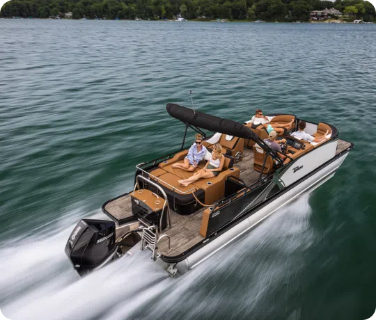 A motorized pontoon boat, fresh from new pontoon boat sales, speeds across a lake with six people on board. The boat features cushioned seating and a black canopy. The water is calm, and the shoreline with trees and houses is visible in the background.