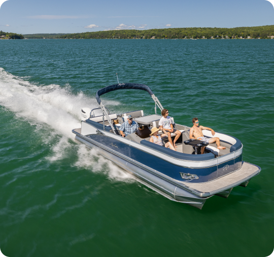 A blue and white pontoon boat cruising on a large, calm lake with four people on board. The boat creates white waves as it moves forward. Behind this scene of new pontoon boat sales is a picturesque shoreline featuring a forest and clear blue sky.