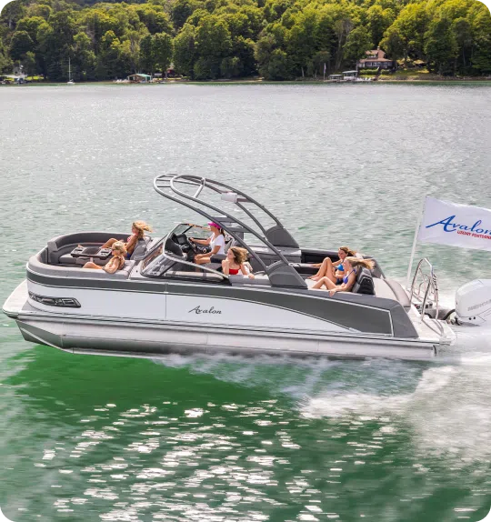 A pontoon boat glides on a lake with six people relaxing onboard, enjoying their recent purchase from new pontoon boat sales. The boat proudly displays an Avalon flag. Trees and houses are visible along the shore in the background. The water is calm and green, making for a serene outing.