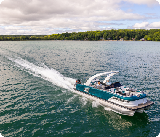 A sleek, modern teal boat speeds across a tranquil lake, leaving a white trail of wake behind it. Several people are onboard, enjoying the ride. The background features a lush, green tree-lined shoreline under a partly cloudy sky—a perfect scene showcasing our new pontoon boat sales.