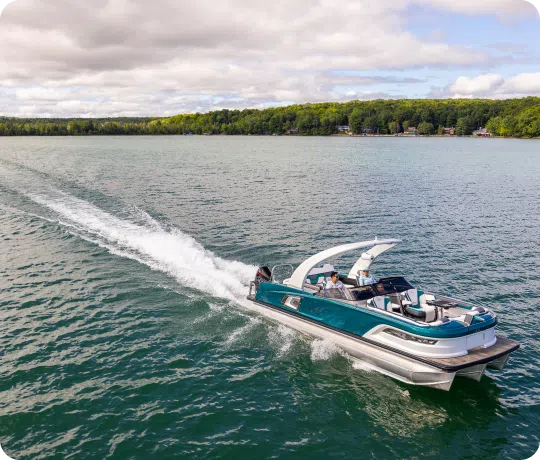 A sleek, modern teal boat speeds across a tranquil lake, leaving a white trail of wake behind it. Several people are onboard, enjoying the ride. The background features a lush, green tree-lined shoreline under a partly cloudy sky—a perfect scene showcasing our new pontoon boat sales.