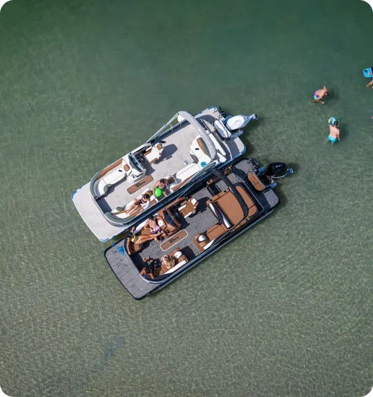 Aerial view of two connected motorboats in clear shallow water, with several people on board and a few others swimming nearby. The sunny, recreational scene highlights the perfect setting for enjoying time on the water, reminiscent of new pontoon boat sales adventures.