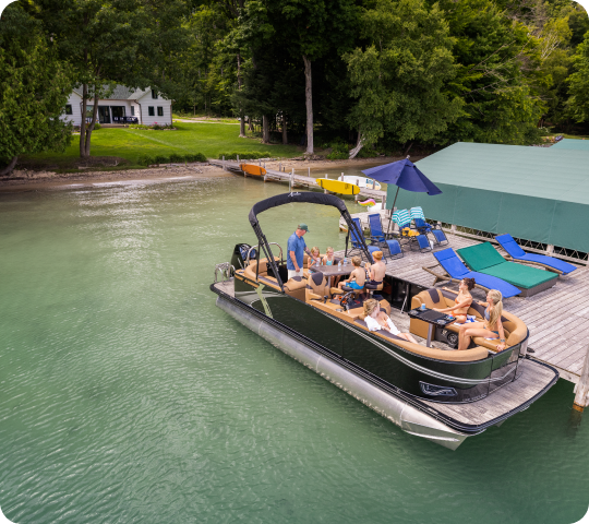 A group of people relaxes on a pontoon boat near a lakeside dock. The dock is equipped with lounge chairs, a hammock, and an umbrella. A house and greenery are visible in the background, creating a serene lakeside setting, perfect for showcasing new pontoon boat sales.