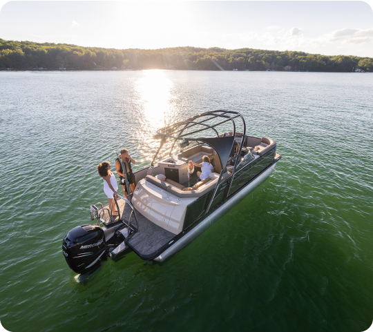 A small group of people enjoy a day on a sleek, modern motorboat on a calm lake with the sun setting over a tree-lined shore in the background. The boat features cushioned seating and is perfect for those considering new pontoon boat sales.