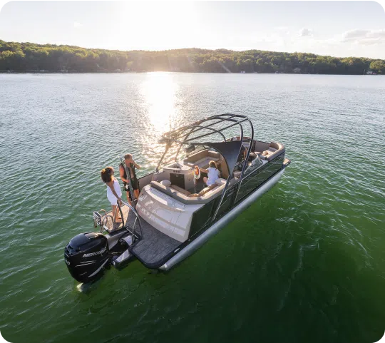 A small group of people enjoy a day on a sleek, modern motorboat on a calm lake with the sun setting over a tree-lined shore in the background. The boat features cushioned seating and is perfect for those considering new pontoon boat sales.