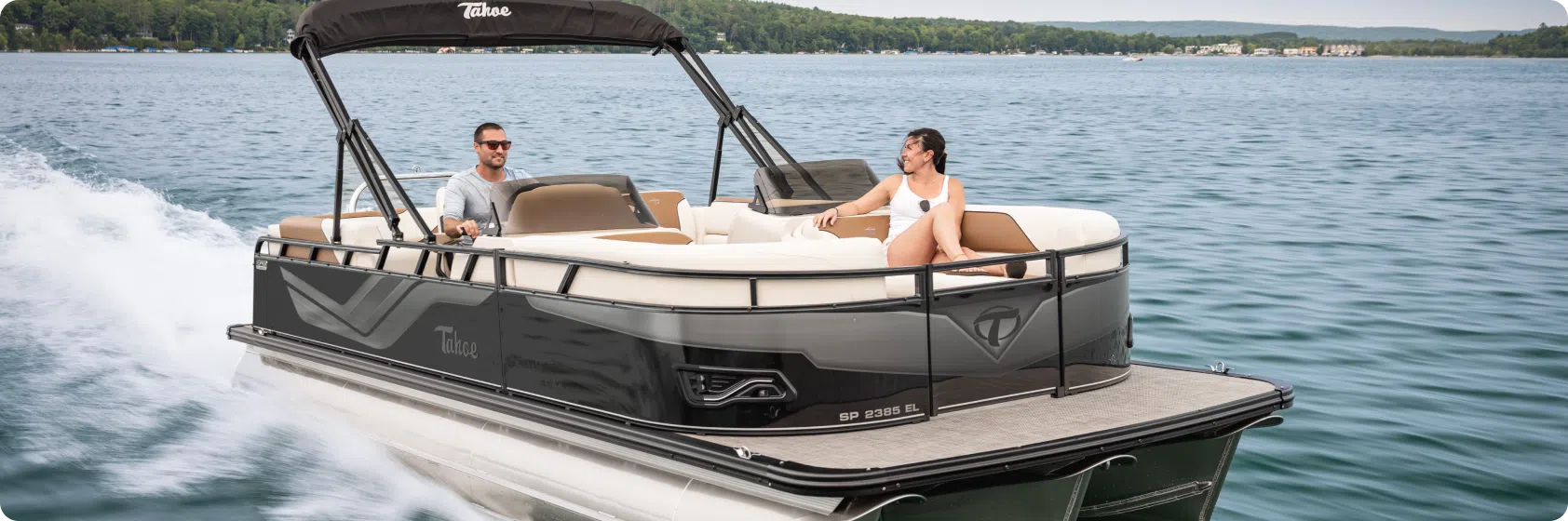 A man and a woman are enjoying a ride on a sleek, modern black pontoon boat with a canopy, gliding across the lake. The boat creates a small wake as it moves through the water with lush, tree-lined shores in the background. They can't help but ponder the cost of such an exquisite pontoon boat.