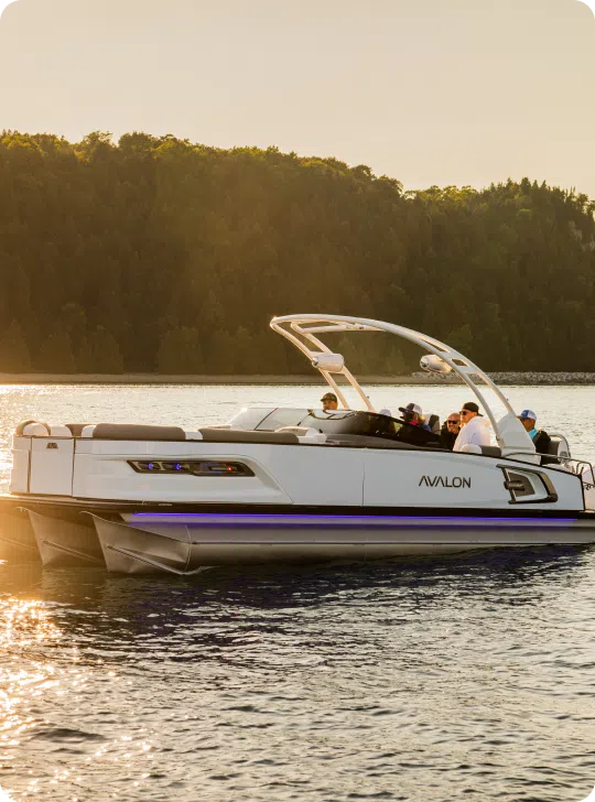 A sleek, modern white boat named Avalon floats on a calm lake during sunset. Several people are on board, enjoying a relaxed outing without worrying about the cost of a pontoon boat. The background features dense, green forested hills with golden sunlight casting a warm glow on the scene.