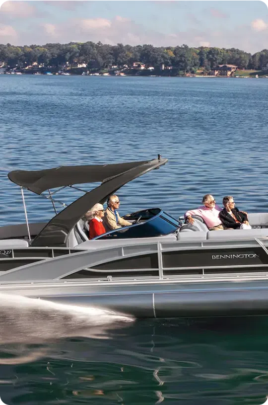 A sleek, modern pontoon boat with a canopy cruises on a calm lake with wooded shores in the background. Four people are seated on the Bennington boat, enjoying the ride. The water reflects the boat and surrounding scenery, showcasing the premium experience that justifies the cost of this pontoon boat.