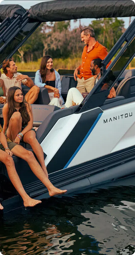 A group of five people enjoys a social gathering on a boat named "MANITOU." Two women and a man sit on the edge with their feet in the water while another man leans against the boat, discussing the cost of pontoon boats with a fifth person standing engaged in conversation. The scene is relaxed and cheerful.