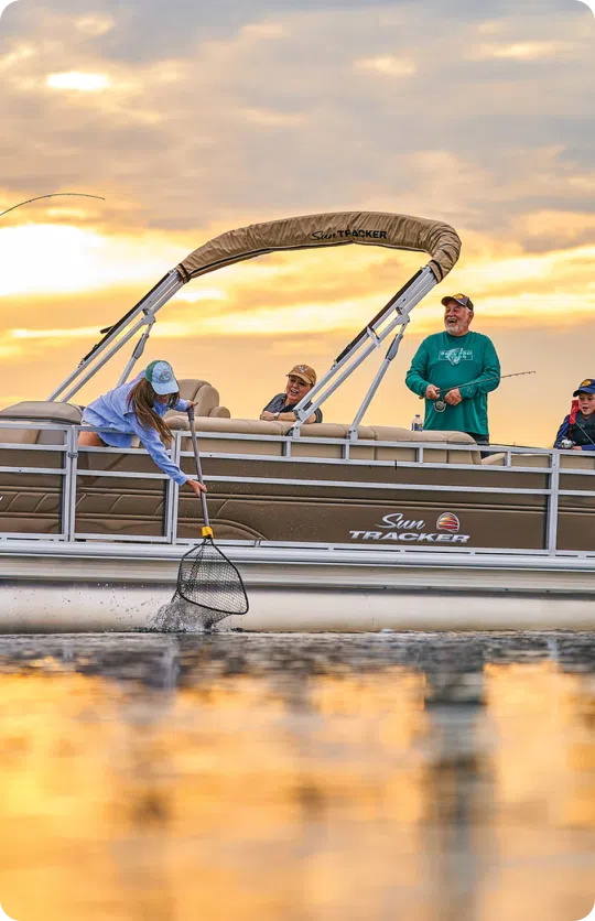 A group of people on a Sun Tracker pontoon boat are enjoying a fishing trip at sunset. One person leans over, using a net to catch a fish from the water, while others watch and smile. The sky is painted with warm sunset colors, reflecting the priceless moments shared regardless of the cost of a pontoon boat.