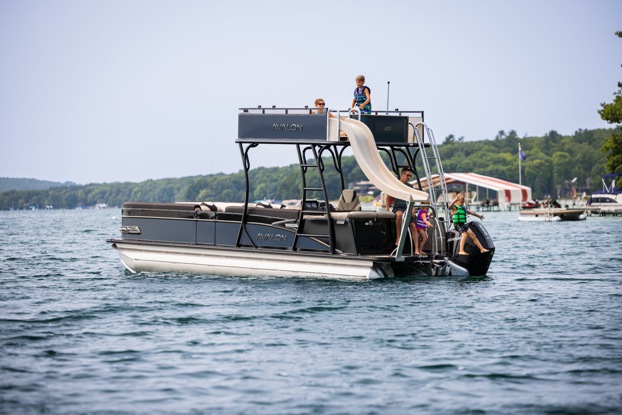 A grey pontoon boat with a slide is on a lake. Children are playing on board; one is climbing the slide ladder while others watch. Trees and buildings are visible in the background. The water is calm and the sky is clear.