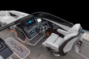 Interior view of a modern boat cockpit featuring a sleek steering wheel, multiple digital screens, and white leather seating on a wood-textured floor. The name "Avalon" is visible on a mat near the step.